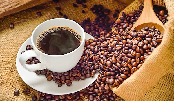 Imagem de uma xícara de café preto sobre um prato branco, ao lado de grãos de café em uma sacola de pano, destacando o aroma e sabor do café.