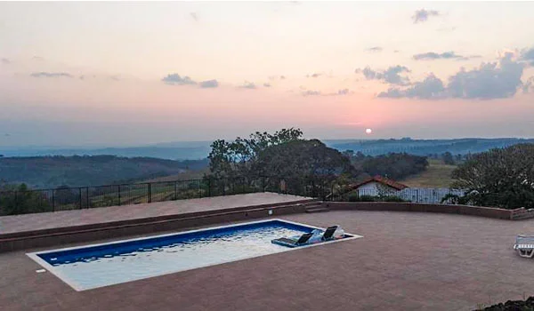 Piscina retangular ao ar livre, com duas espreguiçadeiras dentro da água, localizada em um terraço elevado. Ao fundo, uma paisagem de colinas e vegetação se estende até o horizonte, sob um céu com tons rosados do pôr do sol.