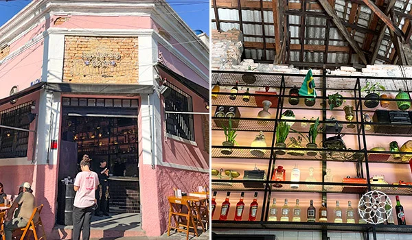 Fachada rosa de um estabelecimento comercial com detalhes brancos e entrada aberta, onde algumas pessoas circulam. Ao lado, o interior apresenta prateleiras metálicas com plantas, vasos, garrafas e objetos decorativos, sob um telhado com estrutura de madeira aparente.