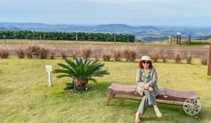 Pessoa sentada em um banco de madeira em um jardim gramado, usando chapéu bege e roupas em tons neutros, com montanhas, plantações e vegetação ao fundo, sob um céu azul parcialmente nublado.