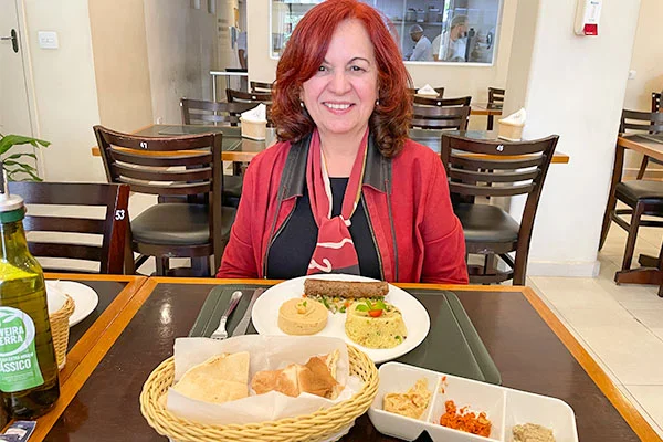 Deusa Rodrigues, sorrindo e sentada à mesa de um restaurante. À sua frente, há um prato com um sanduíche e acompanhamentos, uma cesta de pães e pequenos recipientes com molhos. À esquerda, aparece uma garrafa de azeite. Ao fundo, vê-se o salão com mesas e cadeiras de madeira e uma janela para a cozinha.