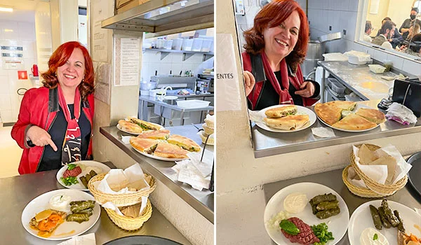 Deusa Rodrigues, sorridente, usando uniforme vermelho e avental preto, aparece atrás do balcão de um restaurante, servindo pratos de comida. À sua frente, há pratos com carnes, salada, legumes e acompanhamentos, além de cestas com pães. Ao fundo, vê-se a cozinha do estabelecimento, com bancadas, utensílios e outros funcionários.