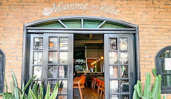 Fachada de tijolos aparentes de um restaurante chamado “Mamma Julia”, com portas duplas de madeira escura e vidro abertas para o interior. Acima da entrada, o nome do restaurante aparece em letras brancas. Vasos com plantas, incluindo espadas-de-são-jorge e cactos, decoram os dois lados da porta.