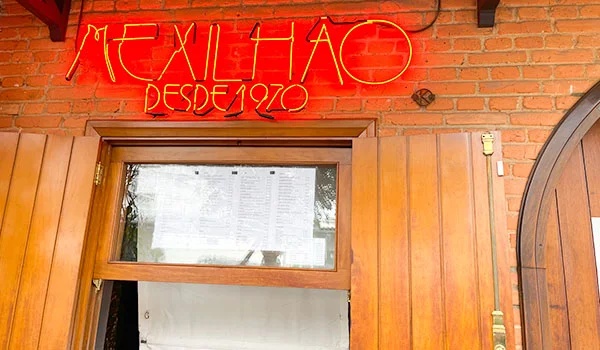 Fachada de um restaurante com parede de tijolos aparentes e porta de madeira. Acima da entrada, uma placa de neon vermelho e amarelo traz o nome “Mexilhão” e a inscrição “Desde 1970”. Na porta há um quadro com o cardápio exposto atrás do vidro.