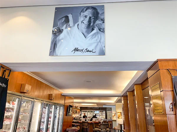 Interior de um restaurante, com paredes e armários revestidos de madeira. No alto, há um quadro em preto e branco com o retrato sorridente de Marcos Bassi e sua assinatura. Ao fundo, vê-se o balcão, mesas e cadeiras, enquanto à esquerda há grandes vitrines refrigeradas com alimentos expostos.