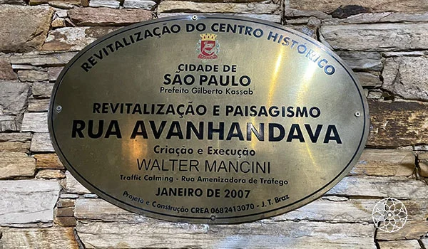 Placa metálica fixada em um muro de pedras, com a inscrição “Revitalização e Paisagismo – Rua Avanhandava”, em São Paulo. O texto informa que a obra foi criada e executada por Walter Mancini, em janeiro de 2007.