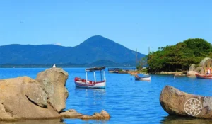 Praia de águas azuis e tranquilas, cercada por grandes rochas, pequenas embarcações e vegetação verde, com montanhas ao fundo sob um céu azul ensolarado.