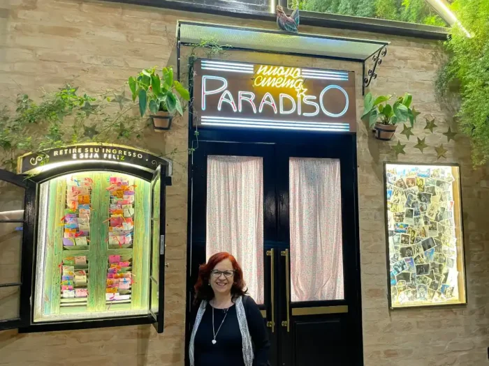 Deusa Rodrigues em frente a uma fachada com letreiro de neon escrito “PARADISO” e entrada de loja com vitrines iluminadas e plantas decorativas na parede de tijolos.