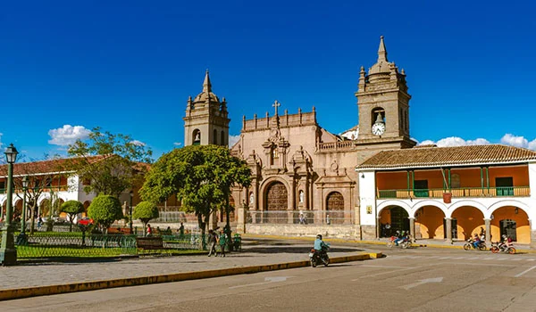 Praça ensolarada com uma imponente igreja histórica de fachada ornamentada e duas torres com relógios, ao fundo. À frente, há um jardim cercado por grades, árvores e bancos. Edificações coloniais com arcadas e varandas contornam a praça, enquanto algumas pessoas circulam pelo local. O céu está azul e sem nuvens Sucre