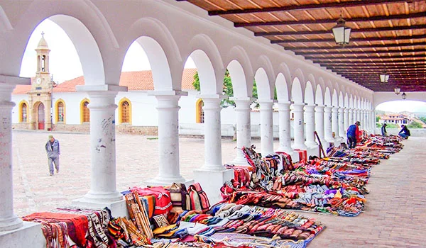 Corredor coberto de um antigo conjunto colonial, com uma longa sequência de arcos e colunas brancas sob um telhado de madeira. À direita, artesanatos e tecidos coloridos estão expostos no chão para venda. Ao fundo, vê-se uma igreja de fachada clara e detalhes amarelos, além de algumas pessoas circulando pelo espaço em Sucre