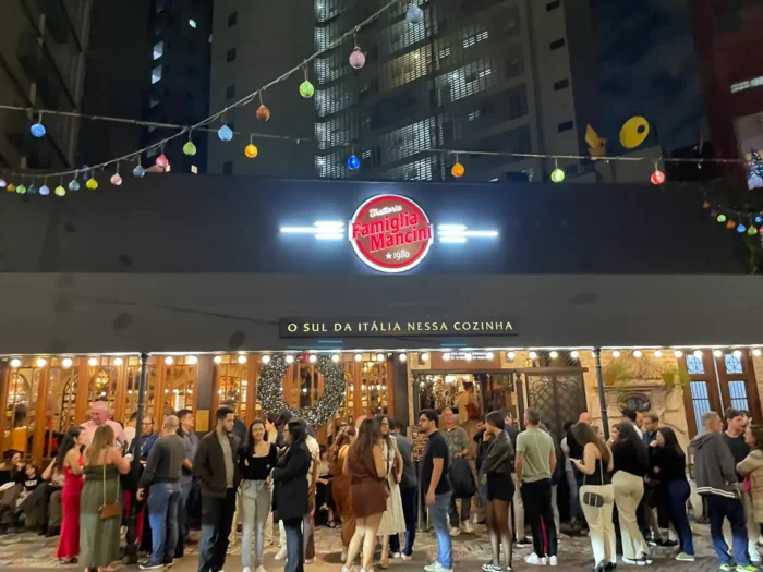 Restaurante com placa “Famiglia Mancini 1980”, área interna decorada e pessoas reunidas em um evento, com luzes e globos pendurados no teto.