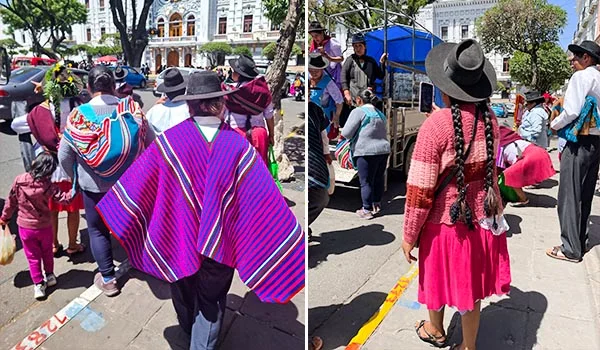 Grupo de pessoas caminhando por uma praça em Sucre, na Bolívia, usando roupas tradicionais andinas, com ponchos coloridos, saias vibrantes, chapéus de abas largas e tranças. Ao fundo, há árvores, pedestres, veículos e edifícios históricos, em um dia ensolarado.