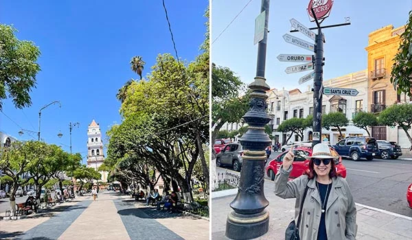 Praça 25 de Maio, em Sucre, na Bolívia. À esquerda, uma alameda arborizada leva à torre de uma igreja sob o céu azul. À direita, Deusa Rodrigues diante de um poste com placas indicativas de cidades bolivianas, cercada por edifícios históricos e carros.