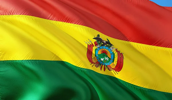 Bandeira da Bolívia hasteada ao vento, com três faixas horizontais nas cores vermelho, amarelo e verde, e o brasão nacional centralizado na faixa amarela.