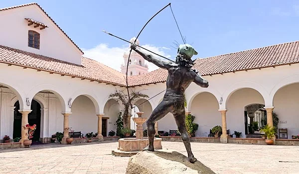 Estátua de um arqueiro indígena no pátio da Casa da Liberdade, em Sucre, Bolívia, cercada por uma construção colonial branca com arcadas, telhados de telhas vermelhas e um pátio pavimentado sob o céu azul.