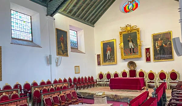 Interior histórico da Casa da Liberdade, em Sucre, Bolívia, com retratos de figuras históricas nas paredes, janelas com vitrais, teto de madeira e fileiras de cadeiras vermelhas com detalhes dourados ao redor de uma mesa central coberta por tecido vermelho.