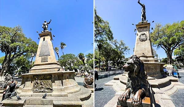 Monumento histórico de piedra ubicado en la Plaza 25 de Mayo, en Sucre, Bolivia, con una estatua de bronce en la parte superior y esculturas de leones en la base, rodeado de árboles y jardines bajo un cielo azul despejado.