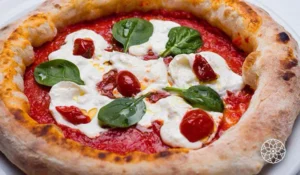 Pizza de massa artesanal com molho de tomate, queijo derretido, tomates-cereja e folhas frescas de manjericão, assada em forno a lenha até a borda ficar dourada e levemente tostada.