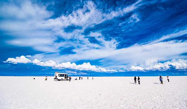 Paisagem do Salar de Uyuni, na Bolívia, com uma extensa superfície branca de sal, céu azul intenso com nuvens e pequenos grupos de turistas ao longe.