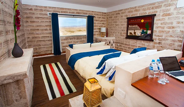 Quarto de hotel com paredes de pedra em tons claros, duas camas de solteiro com roupas de cama brancas e detalhes em azul-marinho, janela com cortinas azuis e vista para uma paisagem desértica. O ambiente conta com piso de madeira, tapete listrado, móveis rústicos, quadro decorativo e bancada com garrafas de água e notebook.
