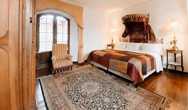 Habitación de hotel de estilo clásico con una cama doble con cabecero de madera tallada, colcha de tonos marrones y rojizos, alfombra ornamental grande en el suelo de madera, sillón junto a una puerta con cortinas y lámparas decorativas a ambos lados de la cama. La estancia tiene una decoración elegante y tradicional.