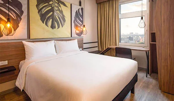 “Habitación de hotel moderna con cama doble amplia, ropa de cama blanca, cabecero de madera, cuadros botánicos en la pared, lámparas colgantes decorativas, cortinas beige, escritorio junto a una ventana con vista urbana y armario de madera al fondo.”