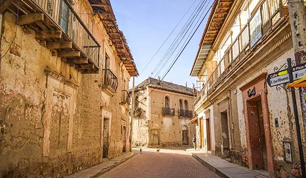 Rua estreita de paralelepípedos em Cochabamba, na Bolívia, cercada por edifícios coloniais de fachadas antigas, paredes desgastadas e varandas de madeira. Fios elétricos cruzam o céu azul, enquanto a luz do sol ilumina a arquitetura histórica da rua. Pontos turísticos da Bolívia.