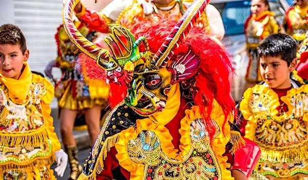Desfile de dança tradicional boliviana, com um dançarino em destaque usando uma fantasia amarela e vermelha ricamente bordada, máscara colorida com chifres ornamentados e uma grande cabeleira vermelha, acompanhado por crianças vestidas com trajes festivos semelhantes.