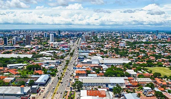 Vista aérea panorâmica de Santa Cruz de la Sierra, na Bolívia, com avenidas movimentadas, edifícios altos, casas, áreas verdes e um céu azul com nuvens brancas. Pontos turísticos da Bolívia.