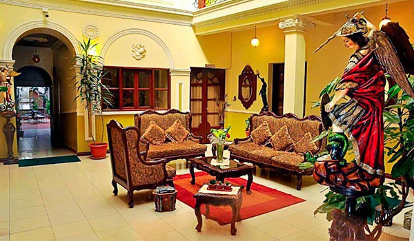 Sala de estar de estilo clásico en un hotel, con sofás y sillones tapizados en tonos marrones, mesas de centro de madera, una alfombra roja y plantas decorativas. El espacio presenta paredes amarillas, arcos ornamentales y puertas de madera. A la derecha destaca una gran escultura decorativa de una figura vestida con ropajes rojos y negros.