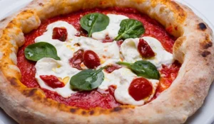 Uma pizza de massa alta e bordas douradas, com molho de tomate, queijo branco cremoso, folhas frescas de manjericão e pedaços de tomate seco. A pizza apresenta algumas áreas levemente tostadas na borda e está servida em um prato branco. O que fazer no Bixiga.