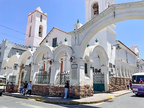 Fachada branca de uma igreja histórica, com arcos ornamentados, detalhes arquitetônicos coloniais, uma torre sineira e muros de pedra na base. Em frente ao edifício, há pedestres, grades de ferro e um ônibus roxo passando pela rua, sob um céu azul e ensolarado.