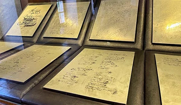 Fotografia de documentos históricos manuscritos, dispostos em vitrines de exposição com molduras escuras e iluminação refletida sobre o vidro. As páginas amareladas apresentam textos escritos à mão, assinaturas e anotações, preservando registros históricos em ambiente de exposição.