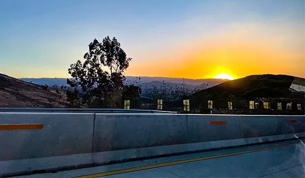 Pôr do sol sobre as montanhas de Sucre, com o céu em tons de azul, amarelo e laranja. A paisagem apresenta colinas escuras, árvores em silhueta e construções com janelas iluminadas. Em primeiro plano, uma barreira de concreto acompanha a estrada, criando uma vista panorâmica da região ao entardecer.