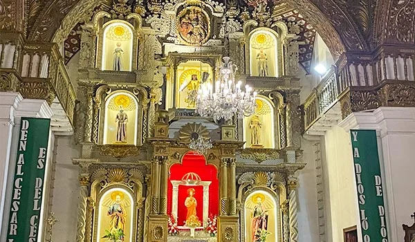 Interior de uma igreja com altar ricamente ornamentado em dourado, imagens de santos em nichos iluminados, uma imagem central de Nossa Senhora sob um dossel vermelho, lustre de cristal e paredes decoradas com detalhes barrocos.