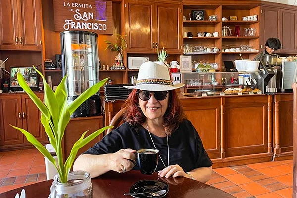 Deusa Rodrigues, usando chapéu branco e óculos escuros, segura uma xícara de café em uma mesa de madeira dentro de uma cafeteria aconchegante, com móveis de madeira, balcão de atendimento, prateleiras com utensílios e uma planta verde ao lado.