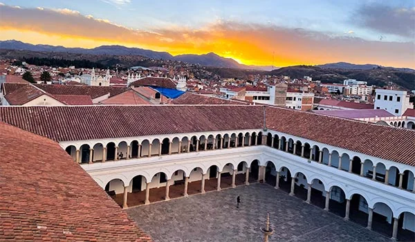 Vista panorâmica de um pátio colonial com arcadas brancas e telhados de telhas avermelhadas, na cidade de Sucre, Bolívia. Ao fundo, casas e edifícios se estendem até as montanhas, sob um céu iluminado pelo pôr do sol em tons de amarelo, laranja e azul.