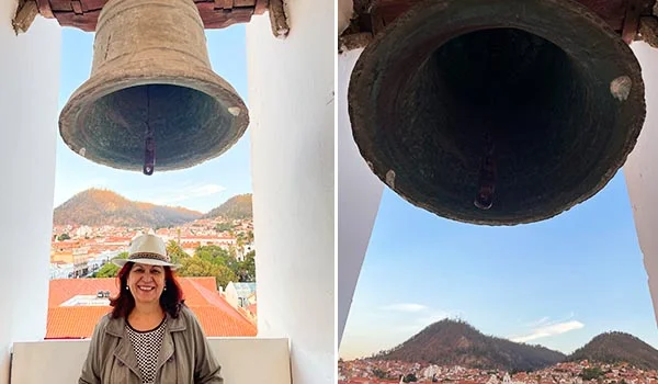 Sino de bronze em primeiro plano, visto por baixo, com uma cidade de telhados avermelhados e montanhas ao fundo. À esquerda, Deusa Rodrigues sorridente usa chapéu claro e casaco cinza, posando sob o sino no mirante de San Miguel, em Sucre, Bolívia.