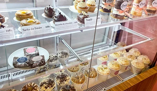Vitrine de uma confeitaria com diversos doces, incluindo tortas de limão, bolos de chocolate, cupcakes, sobremesas em potes e doces decorados, organizados em prateleiras de vidro.
