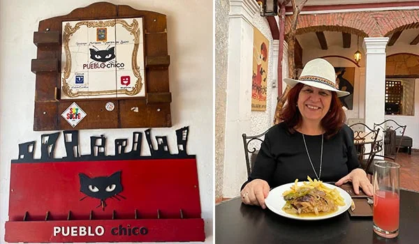 À esquerda, placa decorativa do restaurante Pueblo Chico, com ilustração de um gato preto e detalhes em vermelho, branco e madeira. À direita, uma Deusa Rodrigues, usando chapéu bege e blusa preta, sorri sentada à mesa em um pátio de estilo colonial. À sua frente, há um prato com carne e batatas acompanhado de um copo de bebida avermelhada. O ambiente apresenta arcos de tijolos, colunas brancas e móveis de ferro, criando uma atmosfera acolhedora e tradicional.