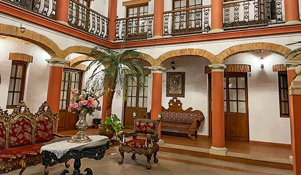 Pátio interno de um edifício histórico, com colunas em tons terracota, arcos, sacadas com grades ornamentadas de ferro, portas e janelas de madeira. O ambiente é decorado com móveis antigos estofados, mesas de centro esculpidas, vasos com flores e plantas tropicais, criando uma atmosfera clássica e acolhedora.