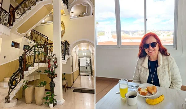 Interior do Hotel San Marino, com uma escadaria elegante de vários lances, corrimãos de ferro trabalhado e vasos com plantas ornamentais. À direita, uma mulher de cabelos ruivos, usando óculos escuros e jaqueta bege, está sentada à mesa durante o café da manhã, com café, suco, pão, queijo e outros alimentos. Ao fundo, uma ampla janela oferece vista para a paisagem urbana sob um céu parcialmente nublado.