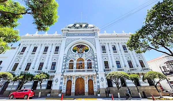 Fachada de um imponente edifício histórico branco, com arquitetura ornamentada, janelas altas, detalhes em relevo e vitrais coloridos na parte central superior. A entrada principal possui grandes portas de madeira e uma varanda com balaustrada. Árvores verdes emolduram o prédio, enquanto carros estacionados e pedestres aparecem na rua, sob um céu azul ensolarado.