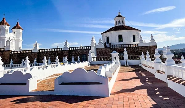 Igreja histórica branca com detalhes barrocos, rodeada por túmulos brancos em um pátio pavimentado com tijolos avermelhados, sob um céu azul e ensolarado.